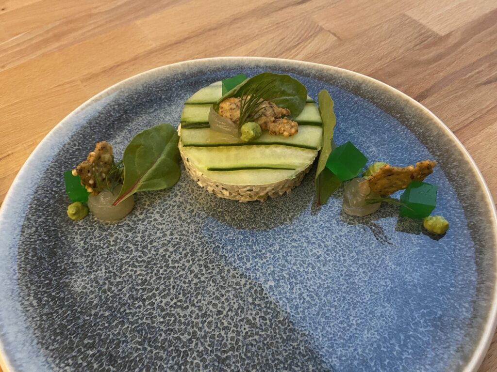 Gurkenscheiben rund ausgeschnitten, passend auf einem flachen Zylinder aus Apfel-Fenchel-Salat liegend, dekoriert mit Blättern, Geleewürfen, Cracker, Cremes und Dillspitzen; im Halbkreis angerichtet auf blauem Teller