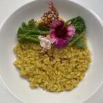 Gold-gelbes Safranrisotto auf einem weißen Teller, dekoriert mit Brokkoli, rosafarbenen Blüten und einem salzigen Cracker
